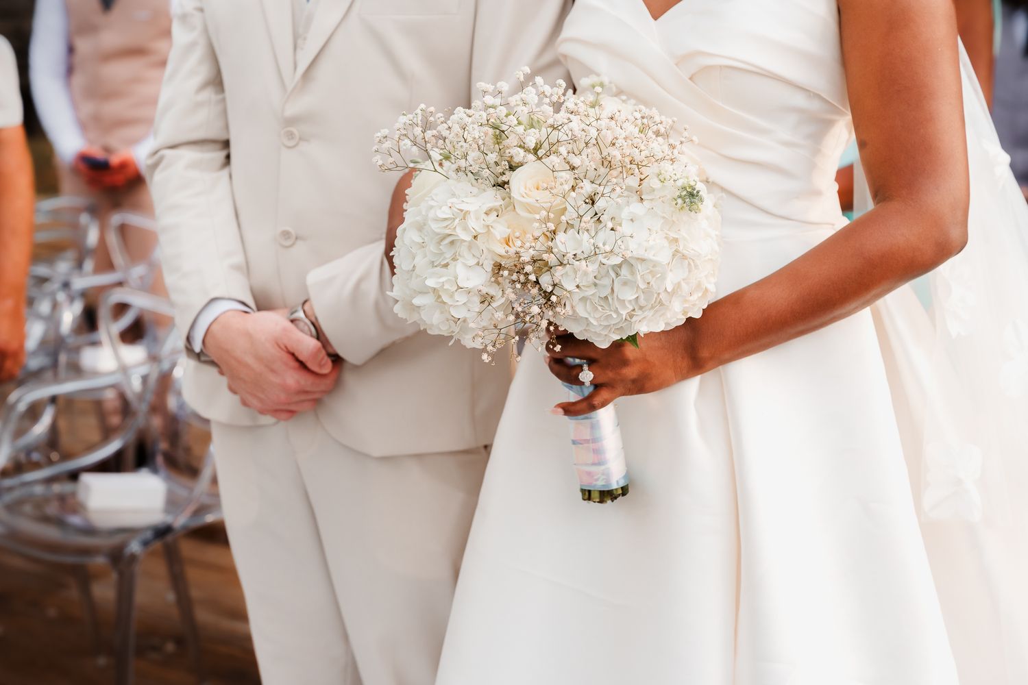 Bride in white gown holding elegant white hydrangea and baby's breath bouquet beside groom in ivory suit.