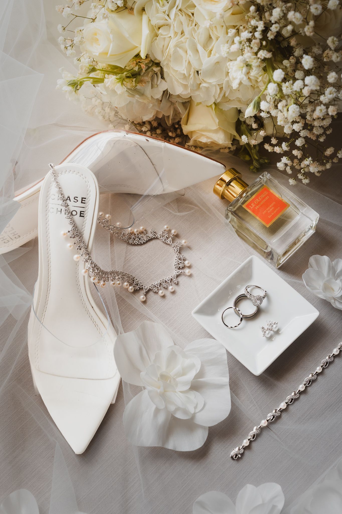 Elegant bridal flat lay with white heels, crystal necklace, rings, perfume bottle, and white floral bouquet on veil.