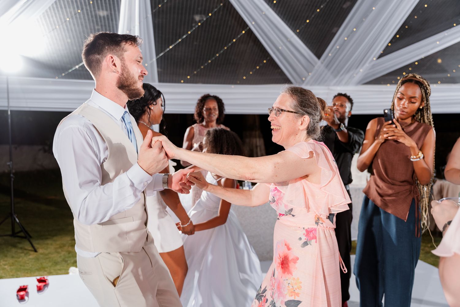 Guests dancing joyfully at an elegant outdoor wedding reception under a white tent with string lights.