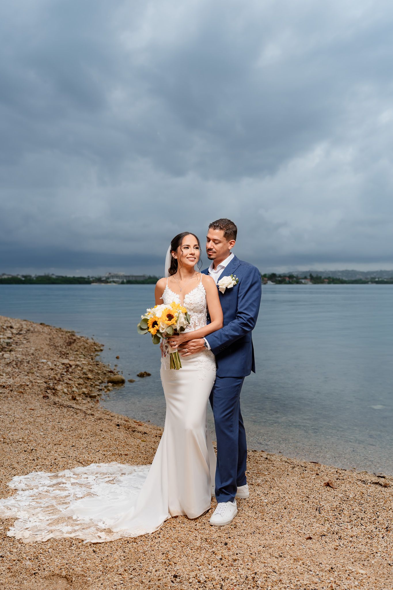 Couple in wedding attire shares intimate moments along a scenic beach shoreline beneath dramatic cloudy skies.