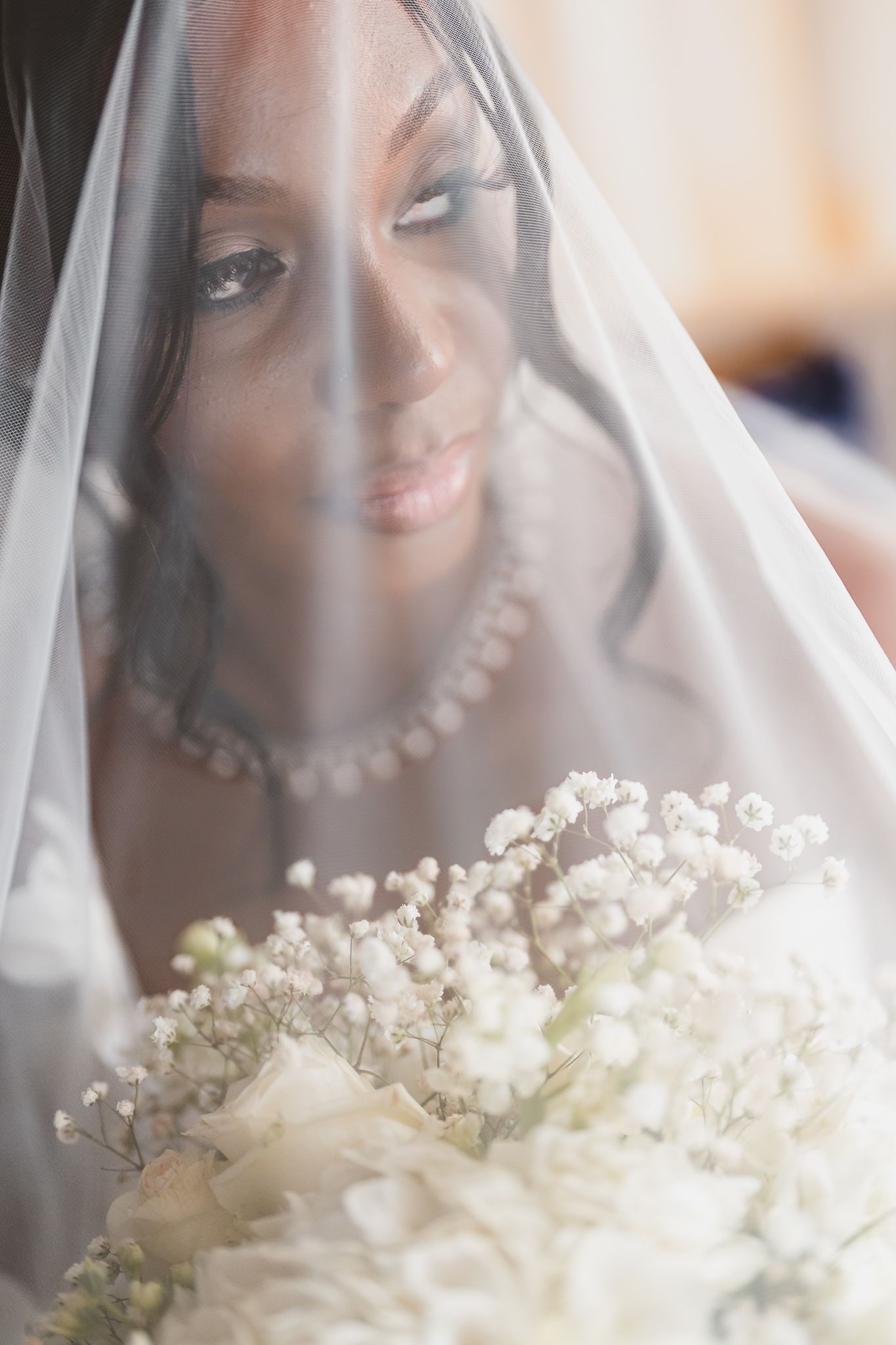 A Black bride gazes down through her white veil, holding a bouquet of white roses and baby's breath on her wedding day.