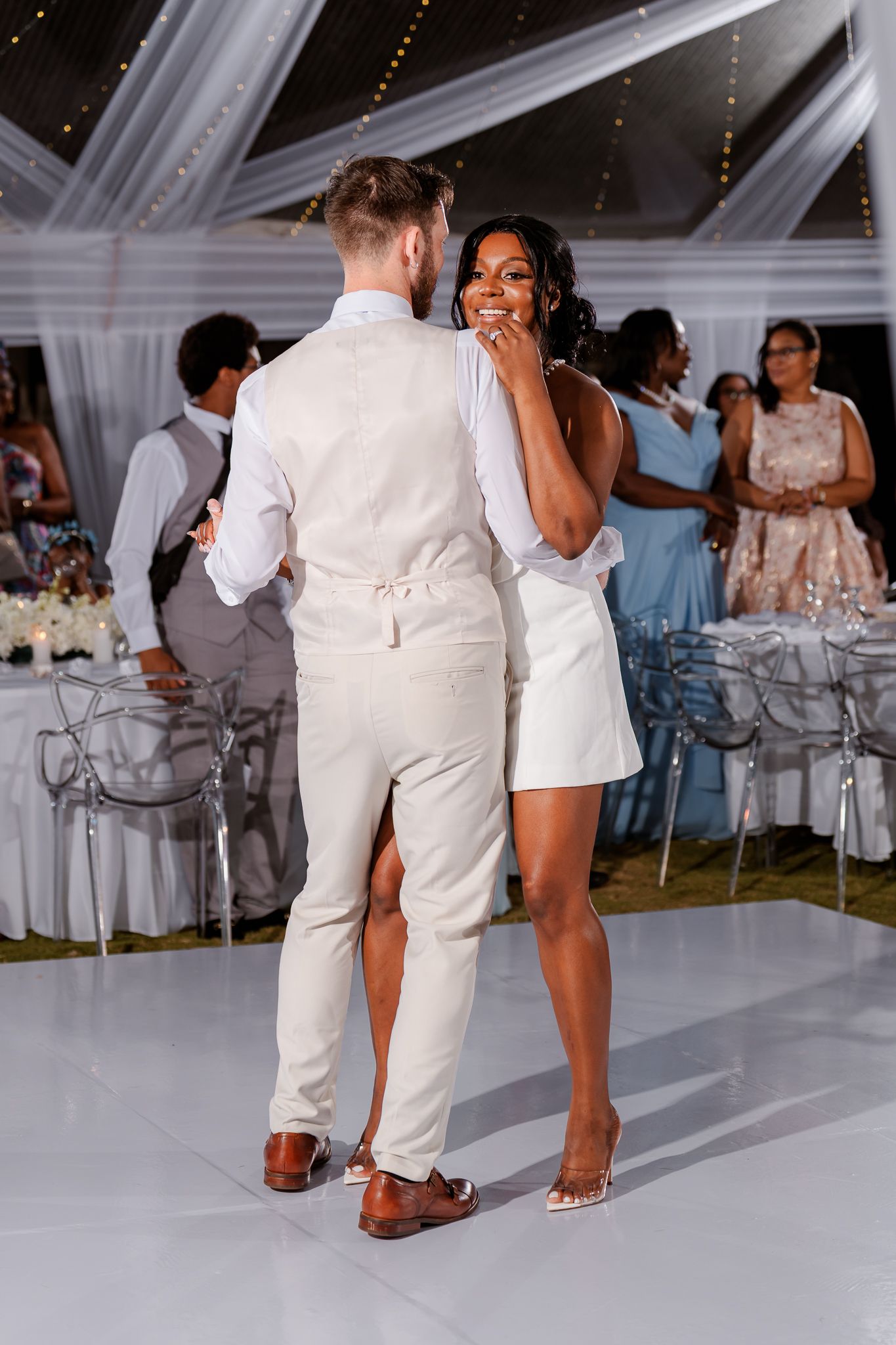 Interracial couple sharing first dance at wedding reception, bride in white mini dress, groom in cream suit, elegant tent venue.