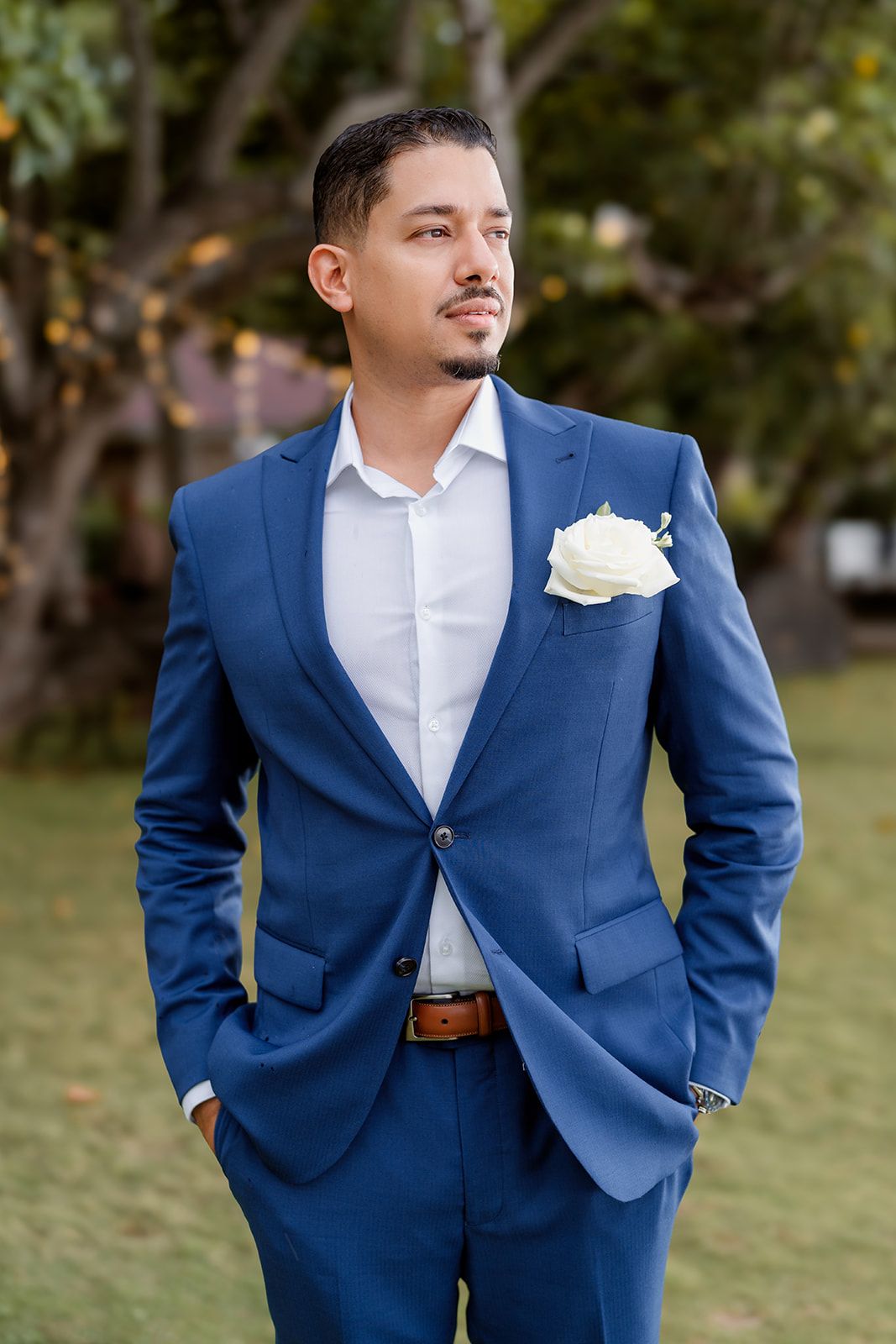 Groom in tailored blue suit with white boutonniere poses outdoors against blurred natural background.