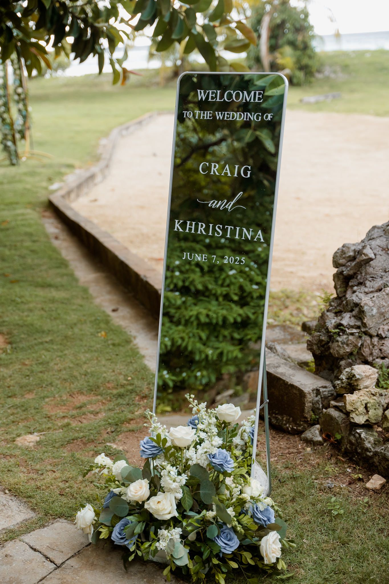 A wedding welcome sign with blue and white floral arrangements sits on a grassy lawn next to stone steps.