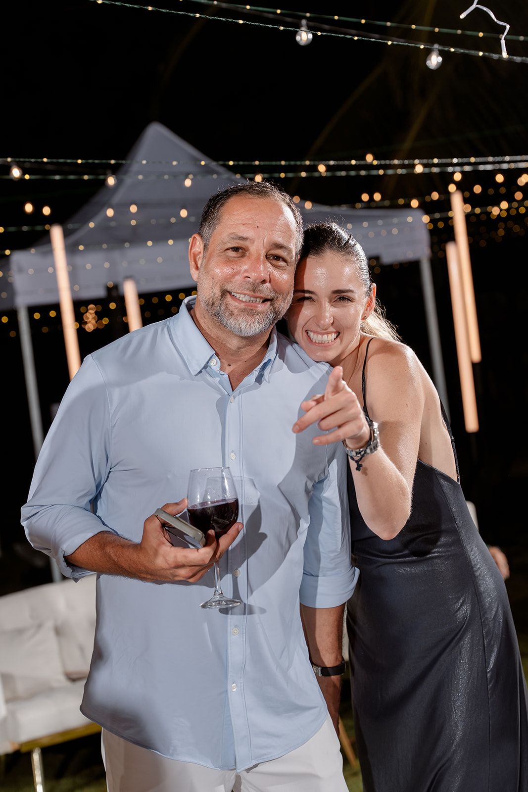 Two people celebrating at an outdoor evening party with string lights and wine under an illuminated tent canopy.
