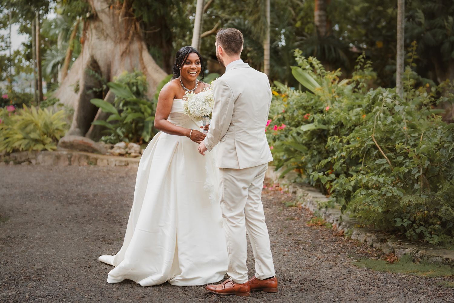 Interracial couple in wedding attire holding hands outdoors, bride in white gown, groom in beige suit, tropical garden.