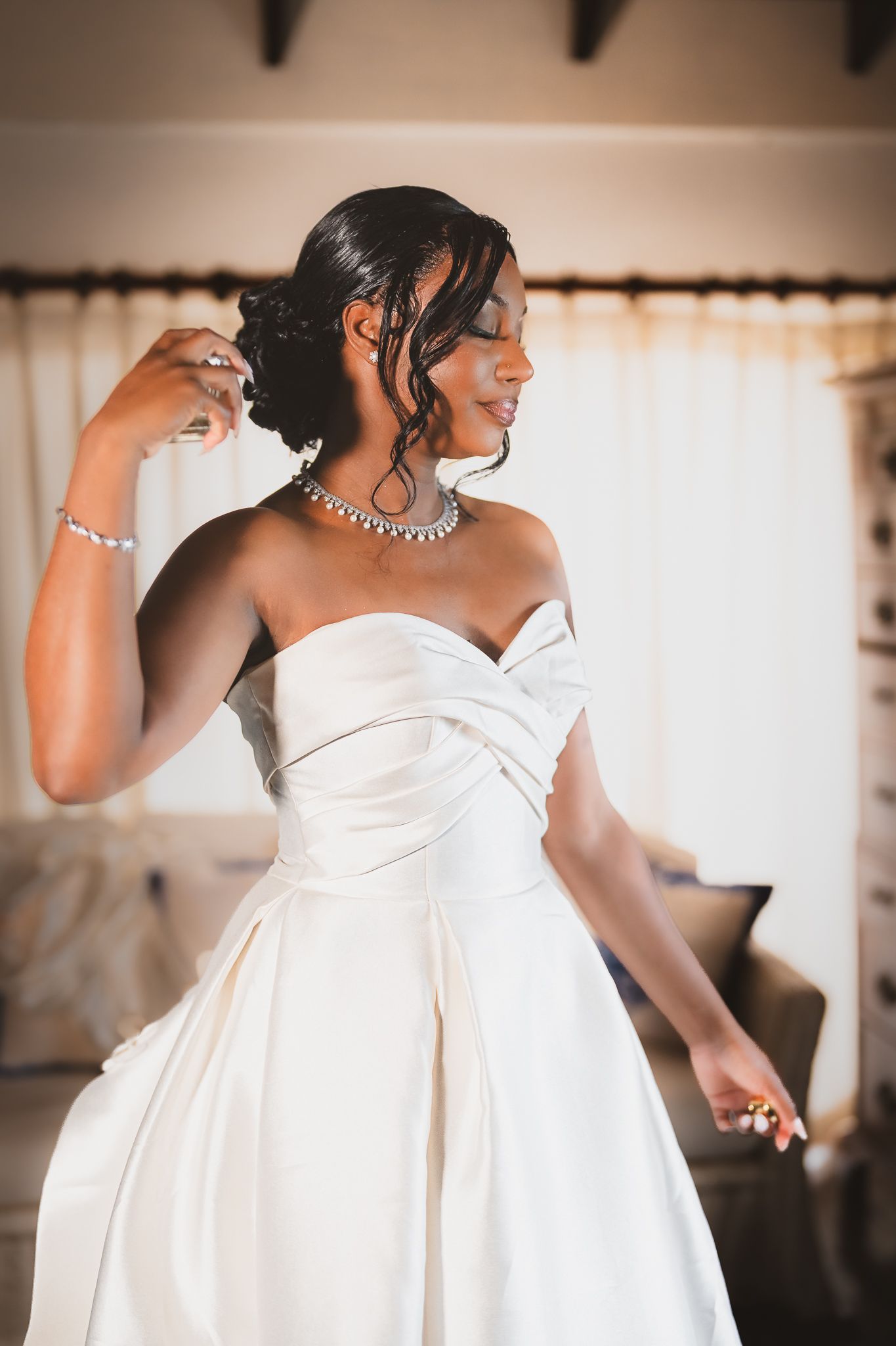 Elegant bride in strapless white satin gown with diamond necklace and bracelet, adjusting her updo hairstyle indoors.