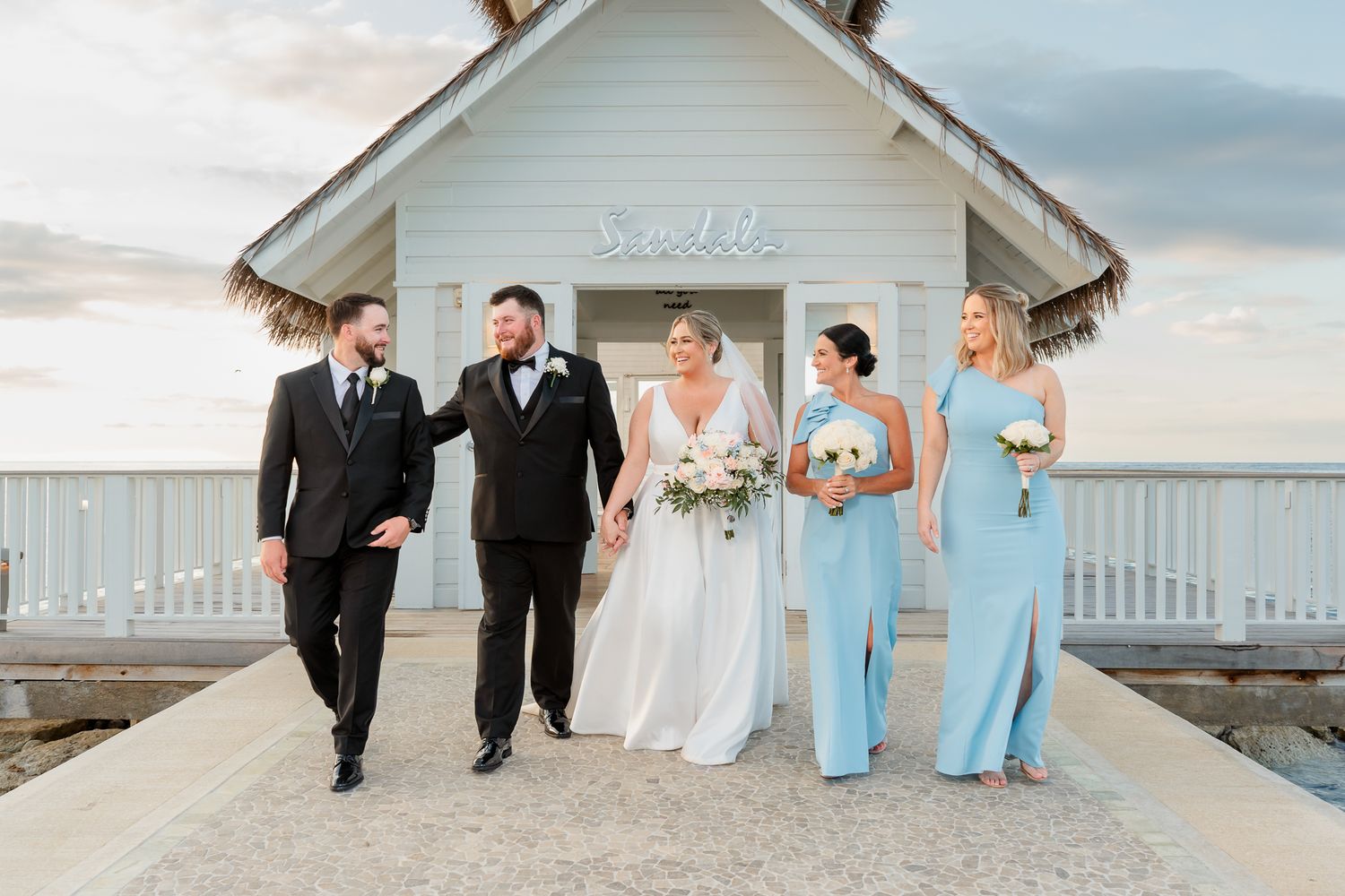 Wedding party walks on pier by white chapel at sunset, bride in white gown, bridesmaids in light blue dresses.