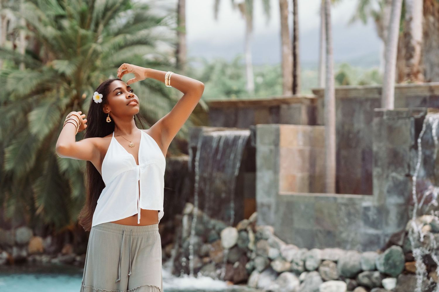 A person in white attire poses elegantly against a tropical backdrop with palm trees and stone walls.