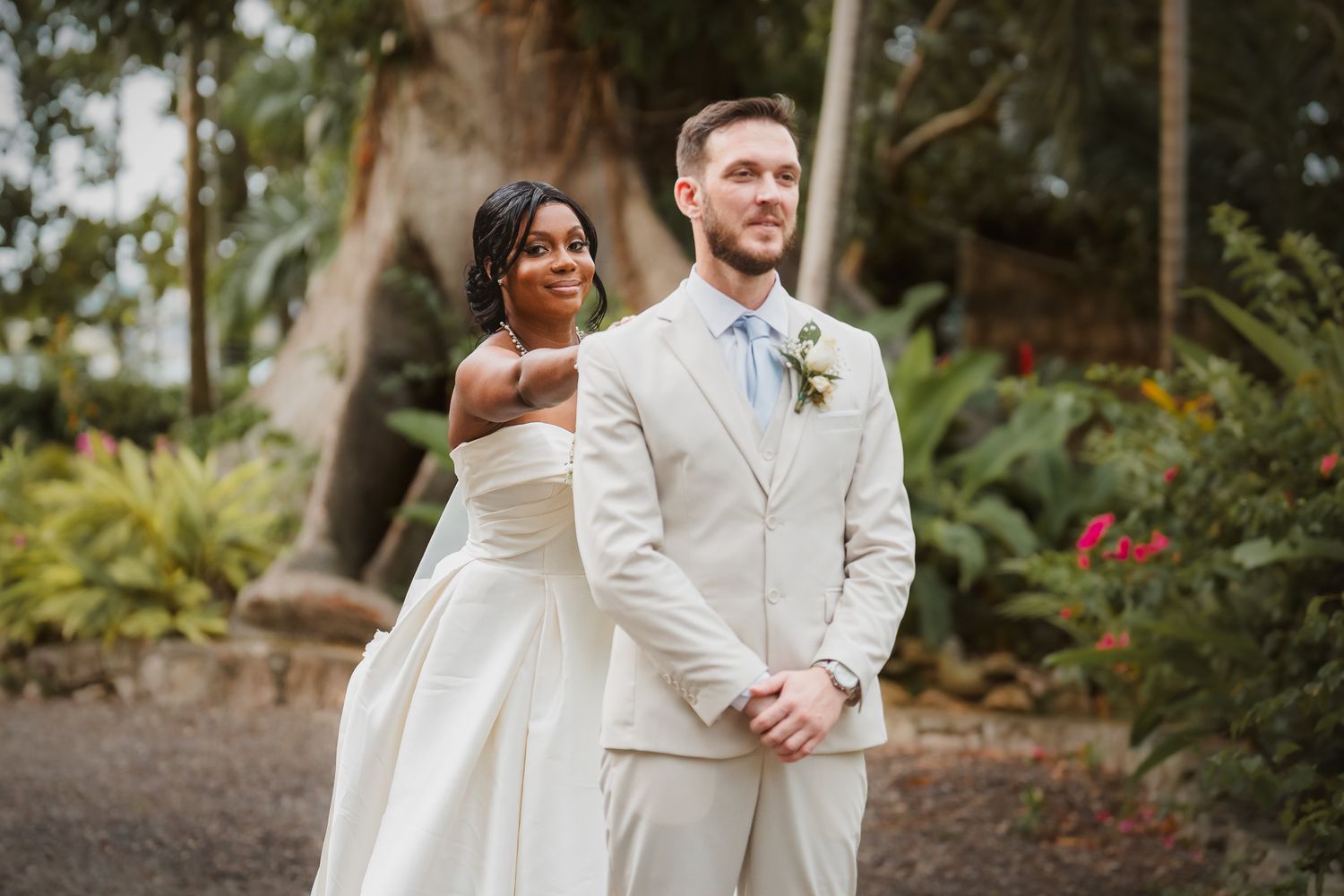 Interracial couple in wedding attire pose outdoors, bride in white gown, groom in cream suit with boutonniere.