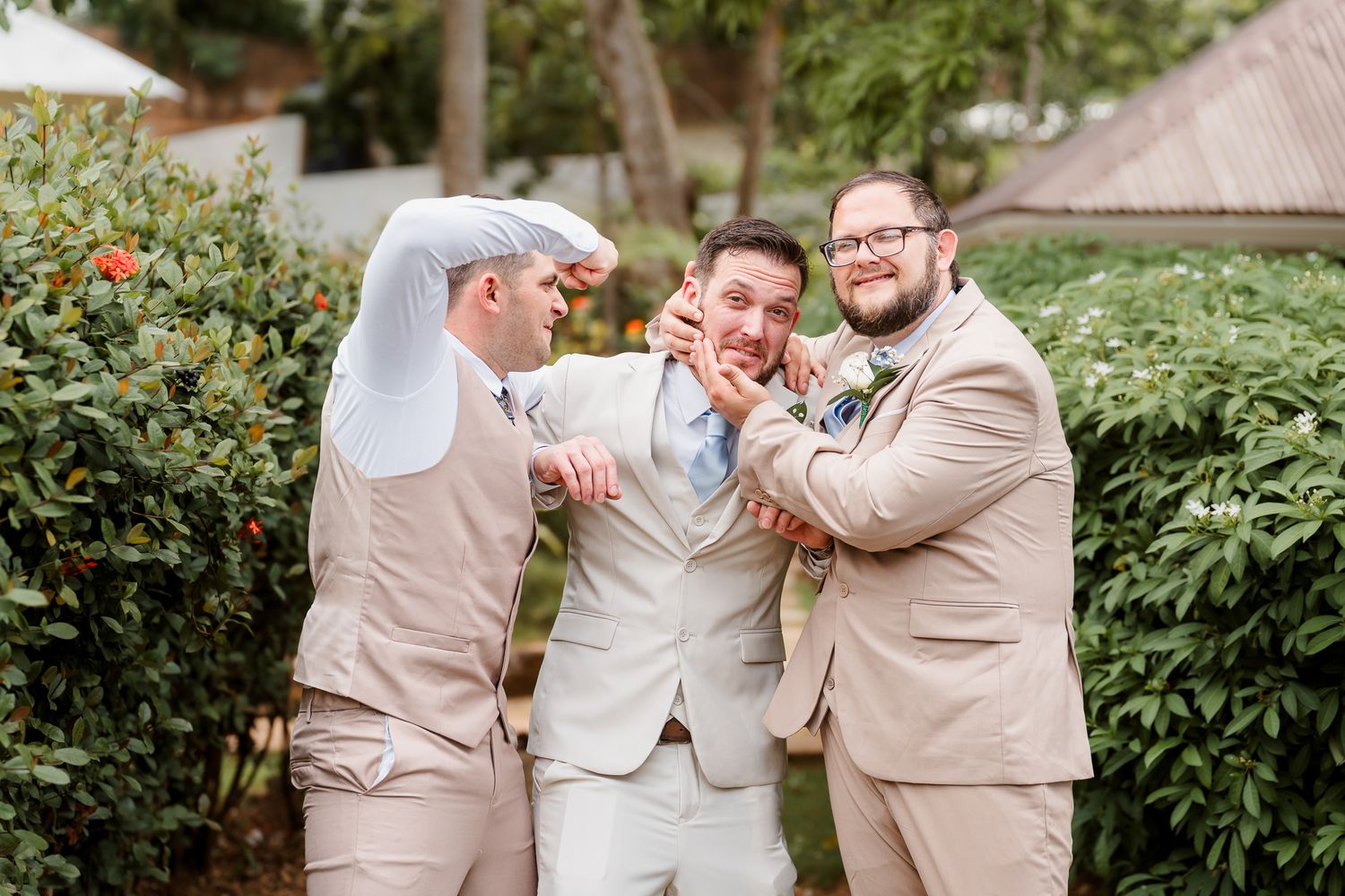 Three groomsmen in tan suits playfully joking around outdoors at a wedding celebration.