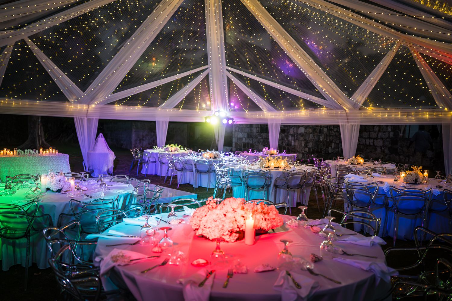 Elegant wedding reception tent with colorful LED lighting, draped white fabric, round tables, and twinkling star ceiling decor.