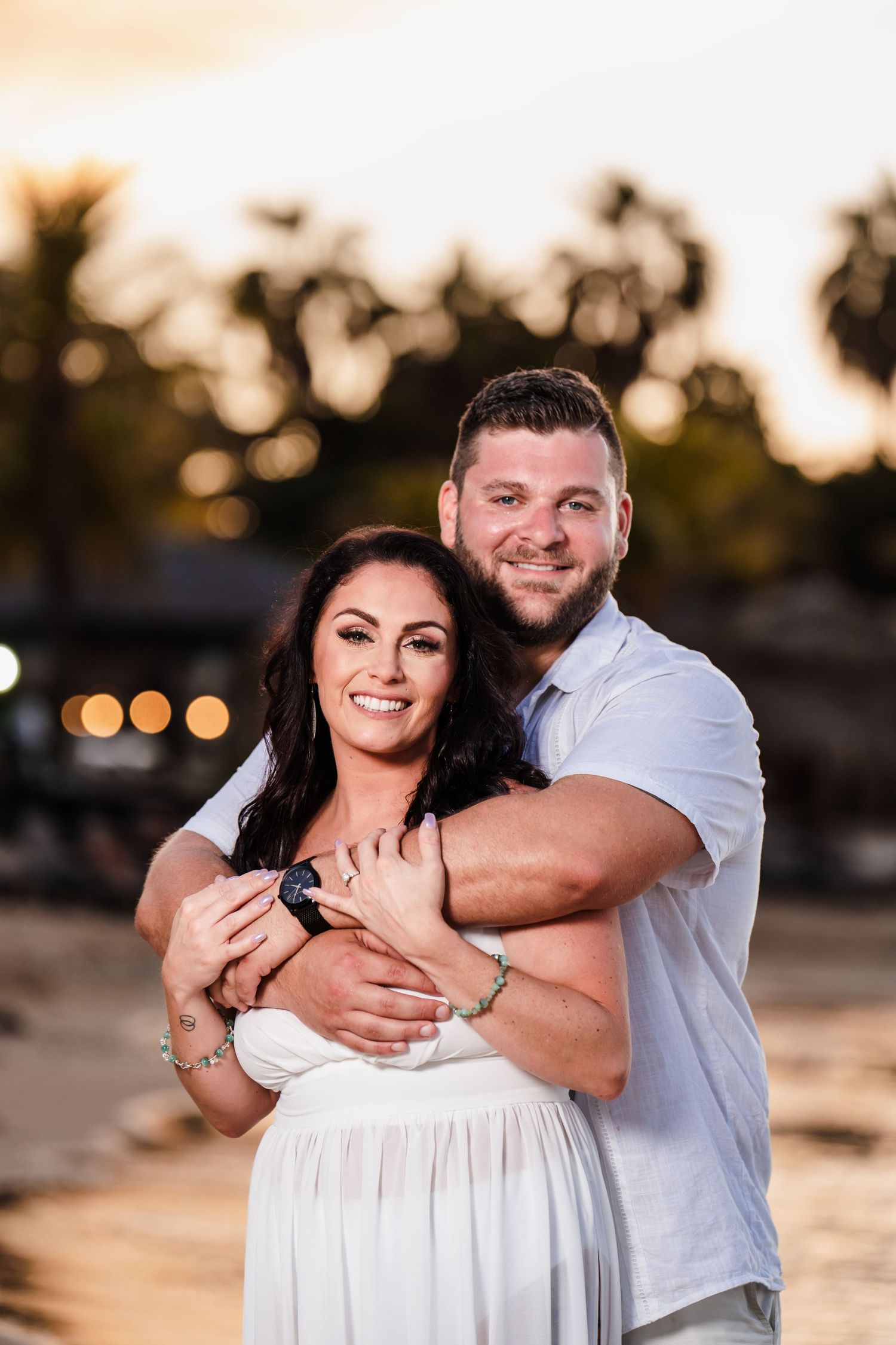 A couple embraces during sunset at a beach, with the person wearing white while their partner holds them from behind.