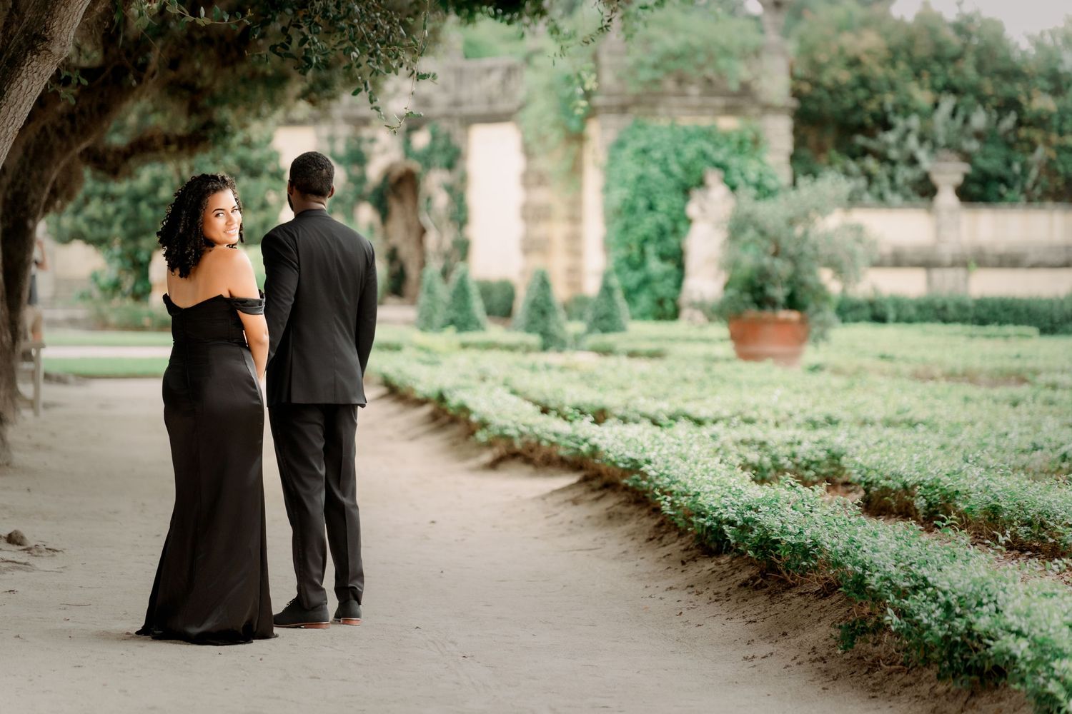 A couple in formal attire walking along a tree-lined garden path during an elegant outdoor event.