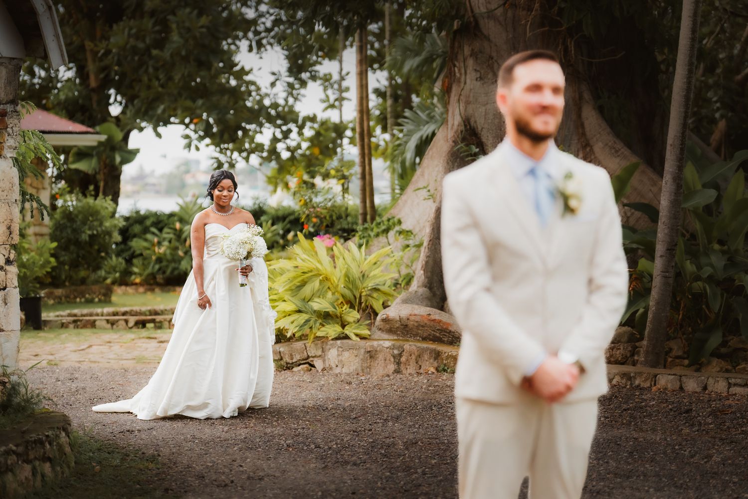 Bride in white gown and groom in tan suit share a romantic first look moment in a lush garden setting.