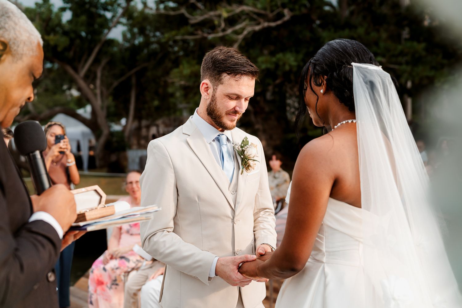 Groom in beige suit holds brides hands during outdoor wedding ceremony while officiant reads from book.