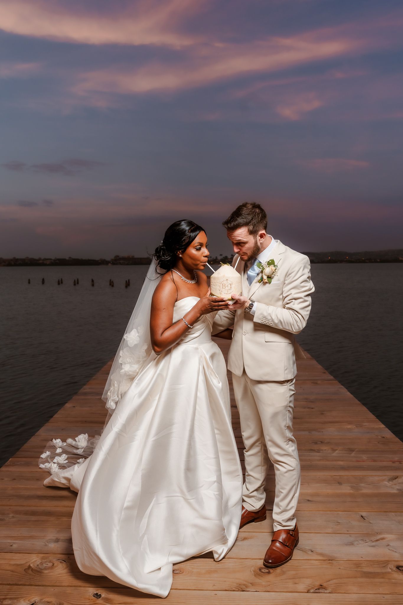 Bride and groom share a romantic sunset moment on a dock, bride in white gown with veil, groom in cream suit.