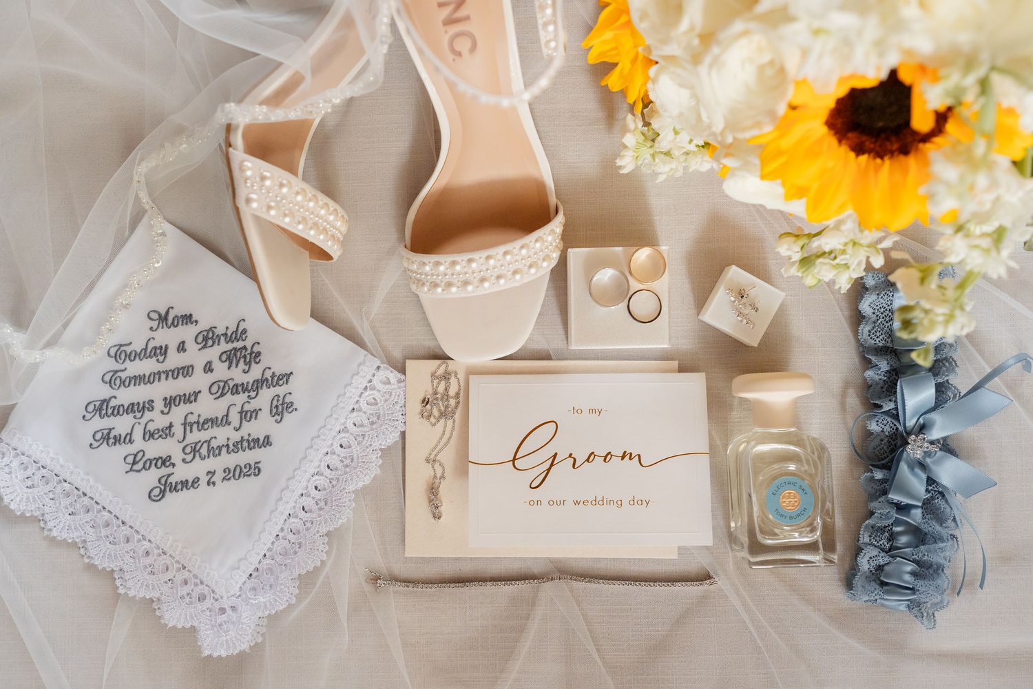 Wedding details flatlay with beige heels, rings, perfume bottle, and sunflowers on white tulle fabric.