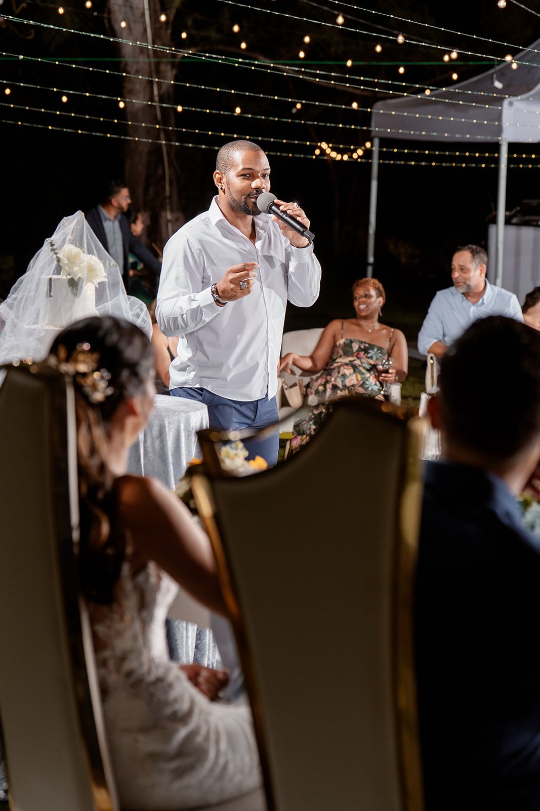 A guest delivers a heartfelt wedding speech under string lights at an outdoor evening reception celebration.