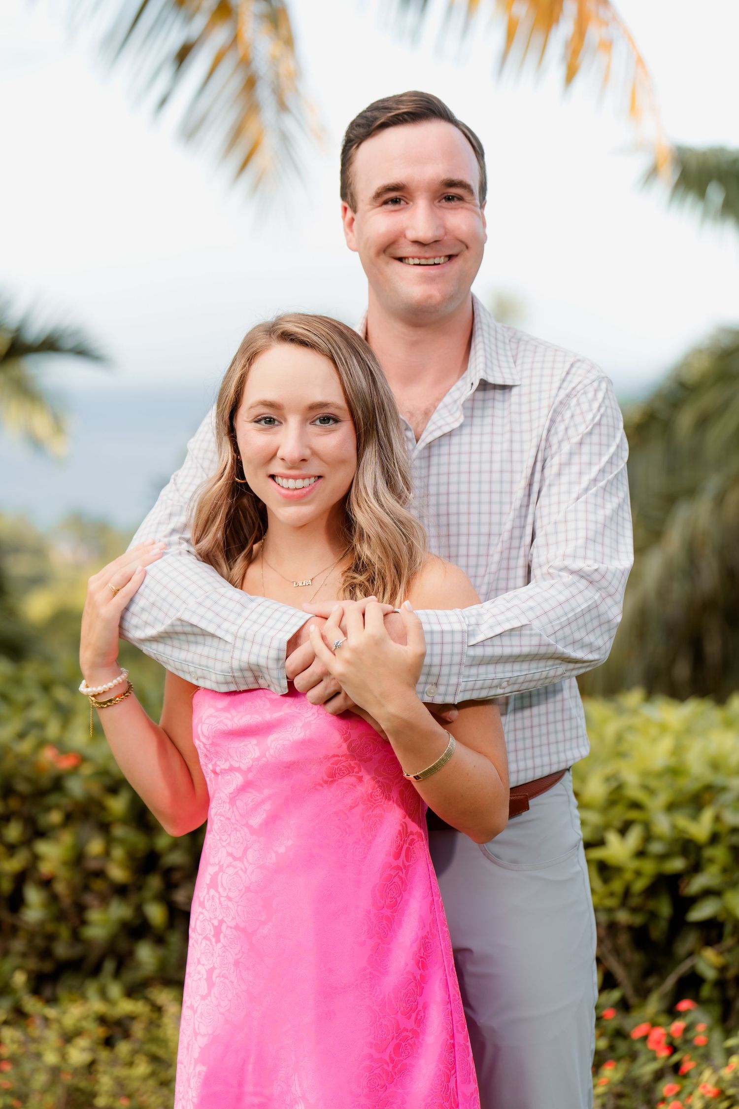 A couple embraces outdoors with palm trees and tropical foliage in the background, she wears a bright pink dress.