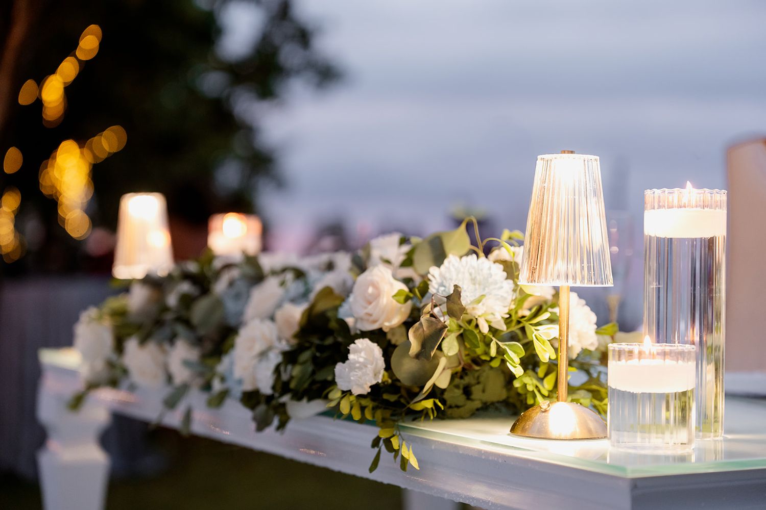 Elegant wedding decor featuring illuminated glass votives and white roses with greenery on a table at dusk.