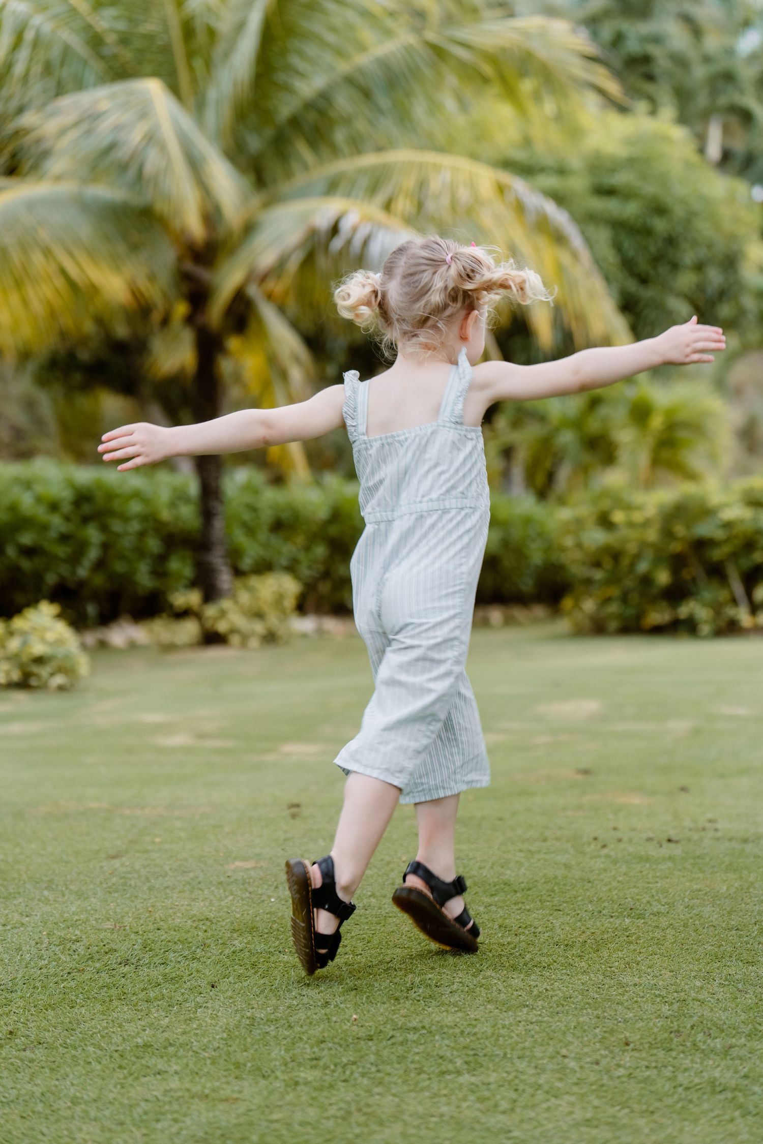 A person in a striped jumpsuit spins joyfully on green grass with palm trees in the tropical background.
