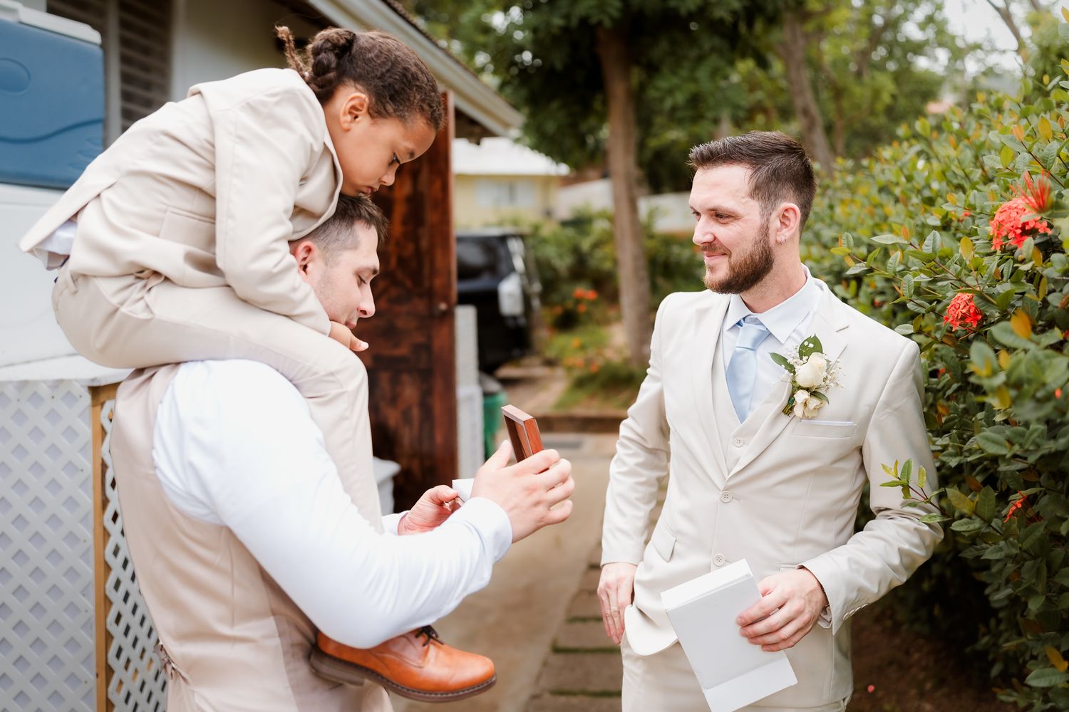 Two groomsmen in beige suits laugh outdoors, one carrying a person on his back, near blooming flowers.