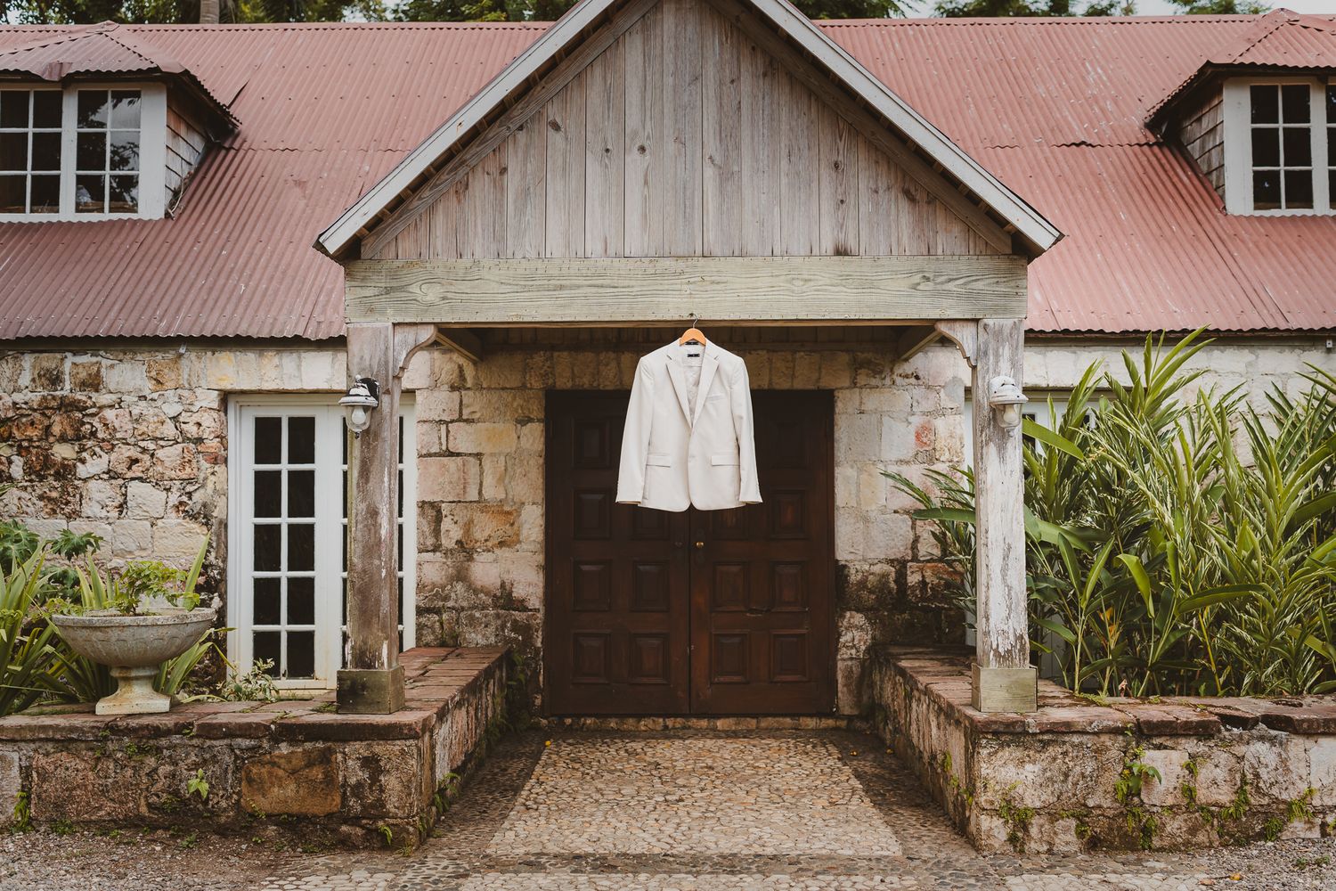 A white blazer hangs above a rustic stone building entrance with wooden doors and red tin roof.