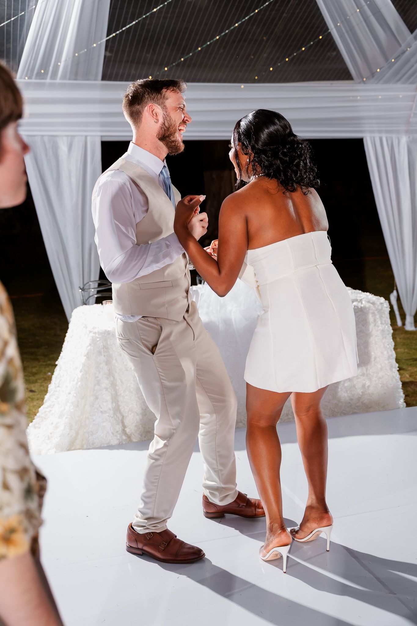 Interracial couple sharing first dance at outdoor wedding reception, groom in tan suit, bride in white mini dress.