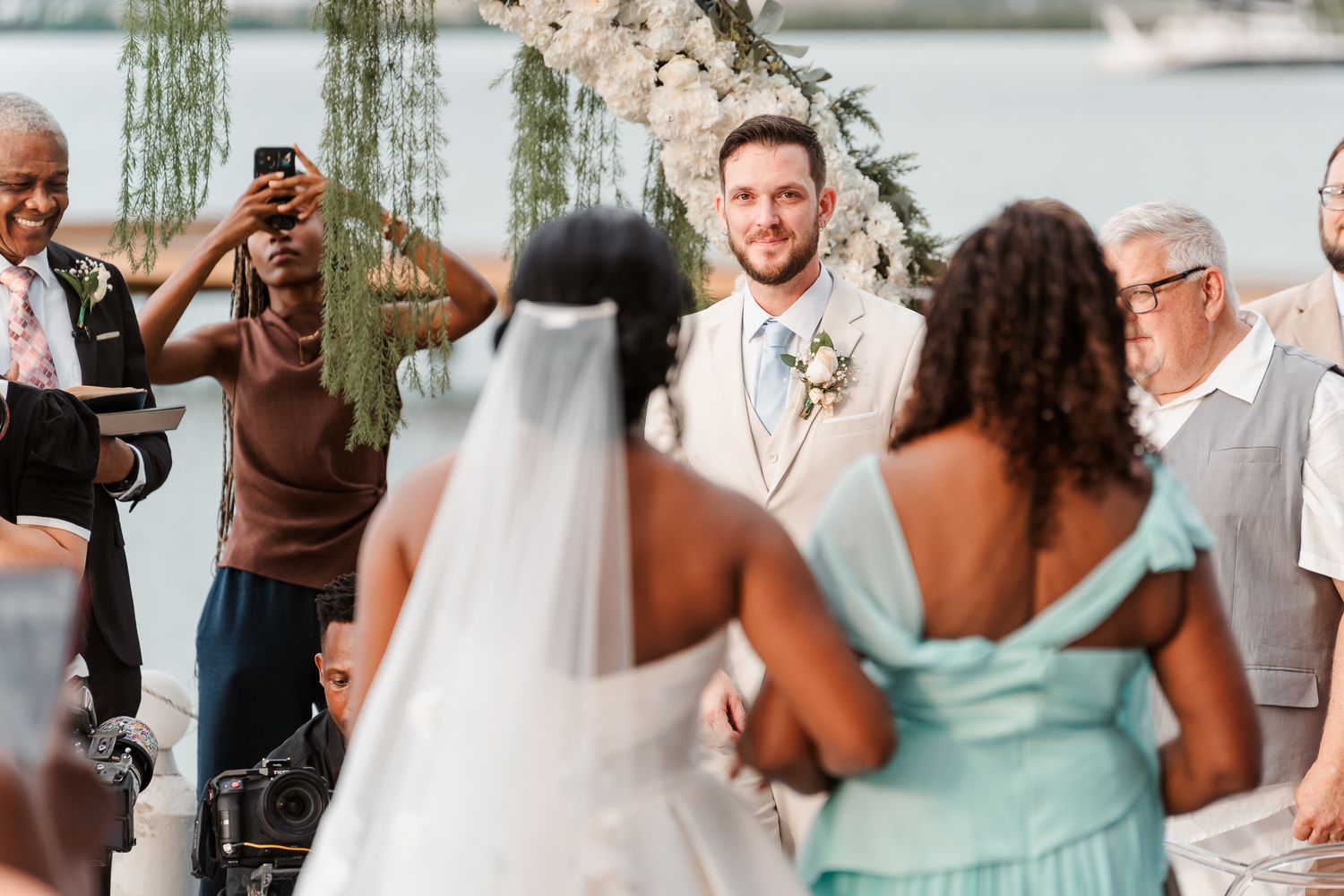 Groom smiling at bride walking down the aisle at an outdoor waterfront wedding ceremony with floral arch.