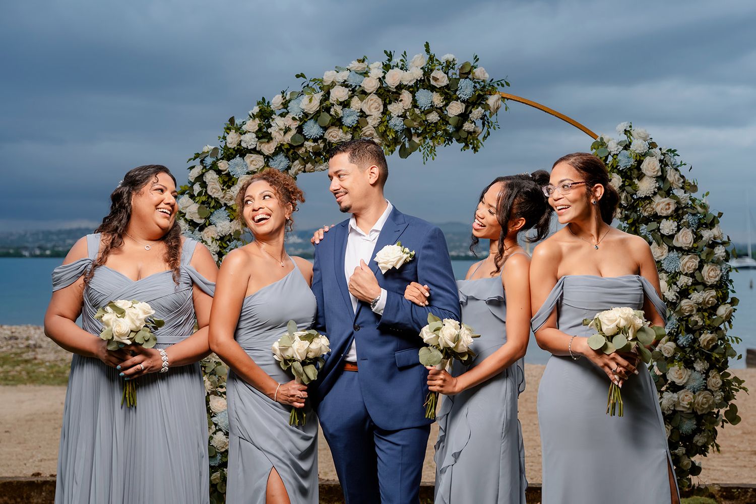 Wedding party shares a candid laughing moment outdoors under a dramatic cloudy sky on the beach.