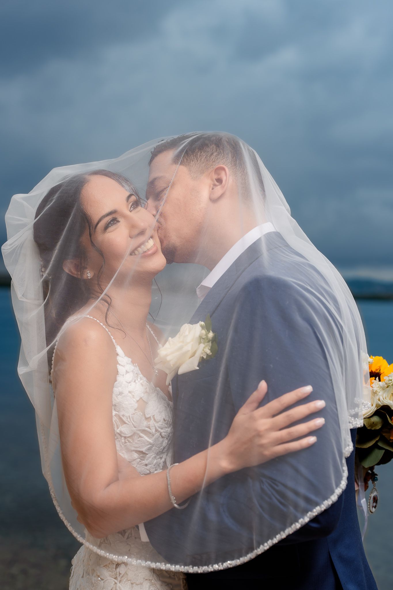 A romantic wedding photograph with a bride in lace dress and flowing veil against a moody blue sky.