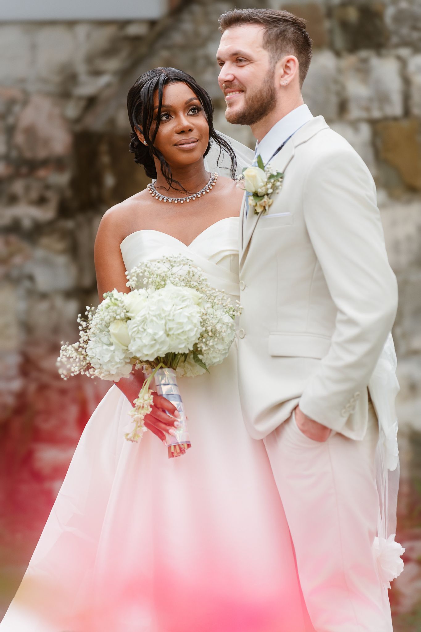 Interracial couple in wedding attire, bride in white ombre gown with bouquet, groom in cream suit with boutonniere.
