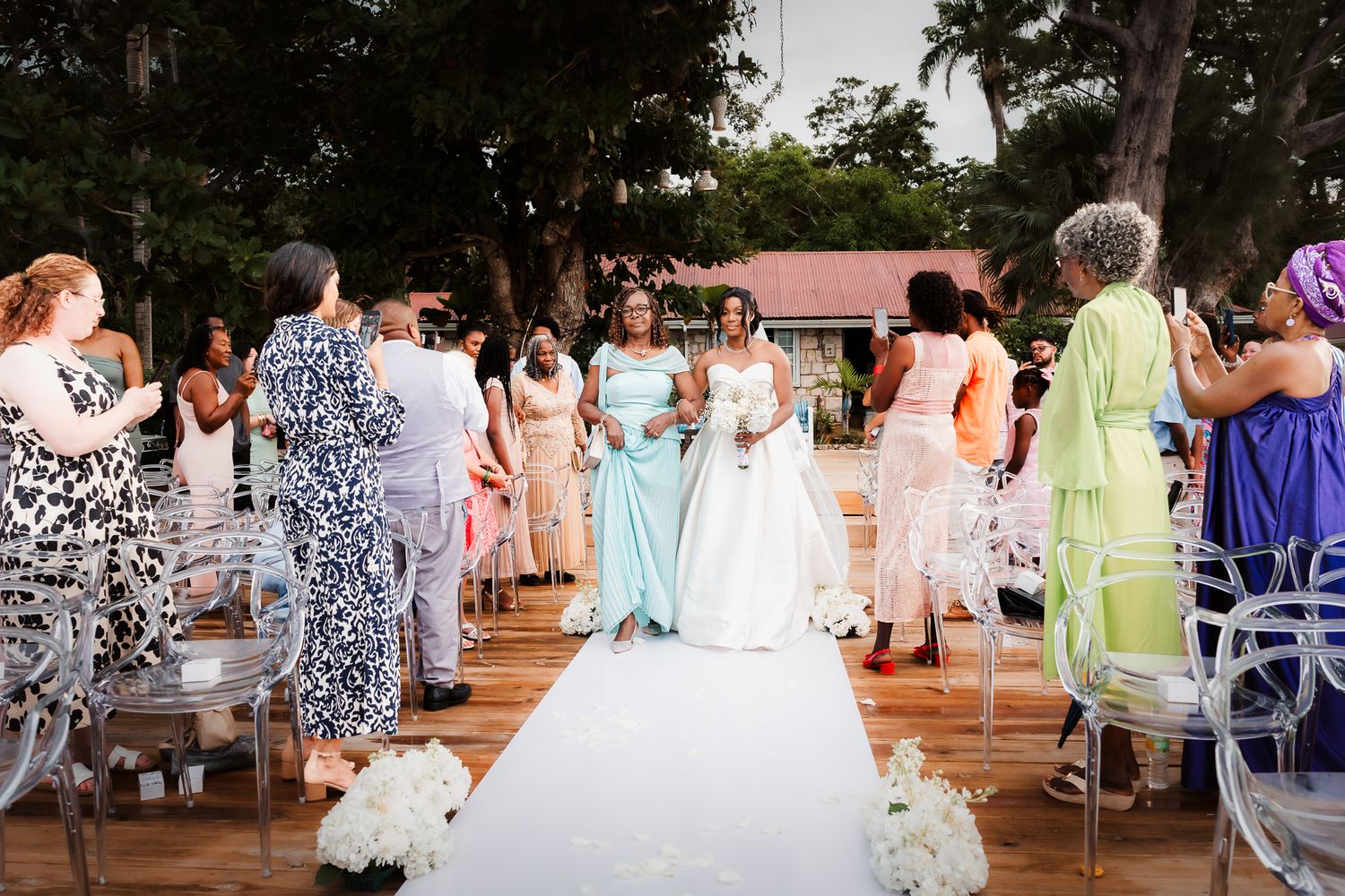 Bride walks down outdoor aisle in white gown with bridesmaids in light blue dresses at elegant garden wedding ceremony.