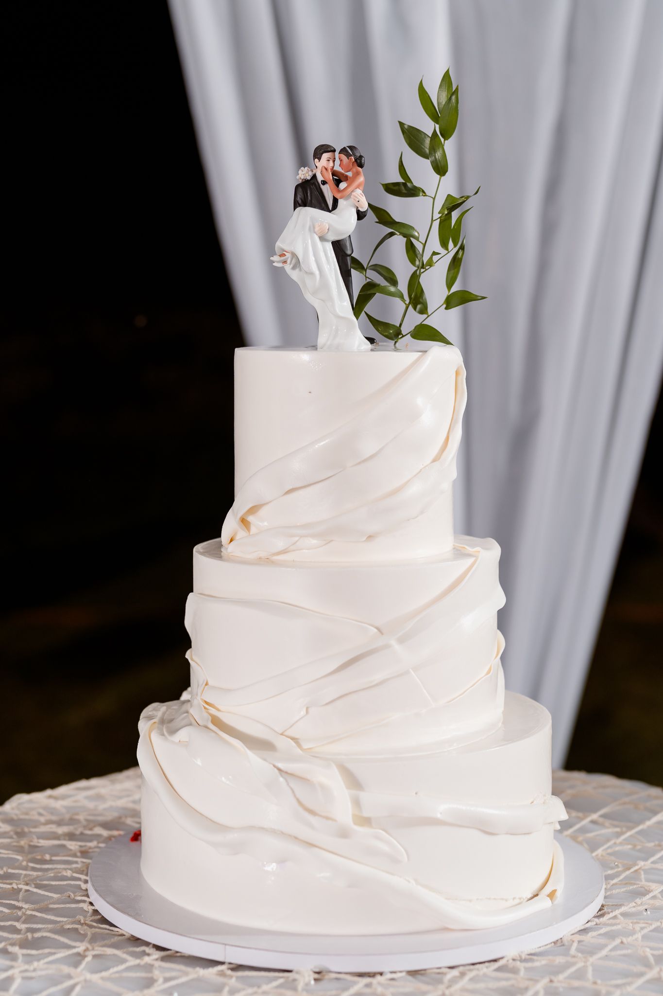 Elegant four-tier white wedding cake with marble design, bride and groom topper, and fresh green leaves.