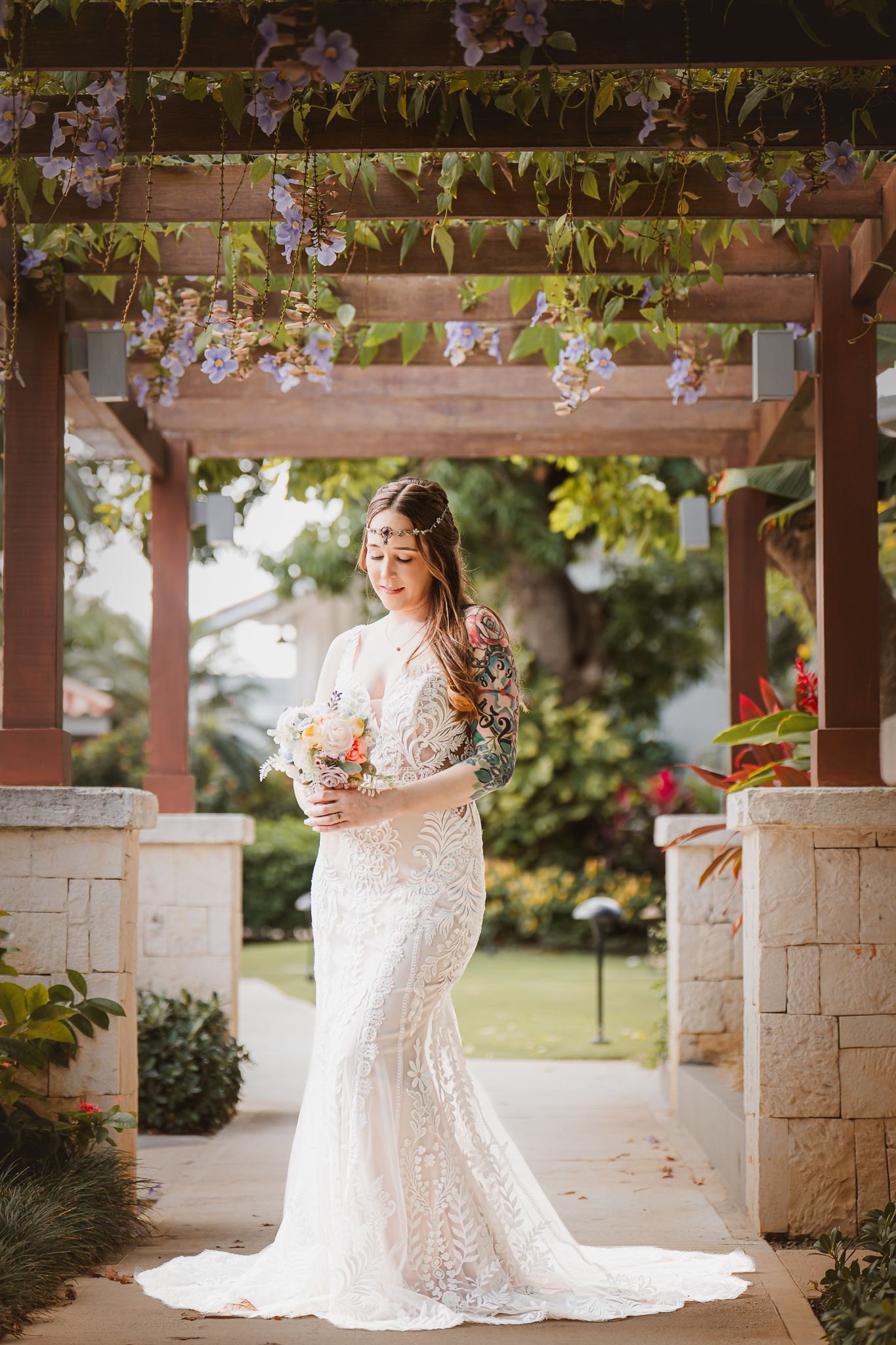 Bride in lace wedding gown holding bouquet under floral pergola in garden.