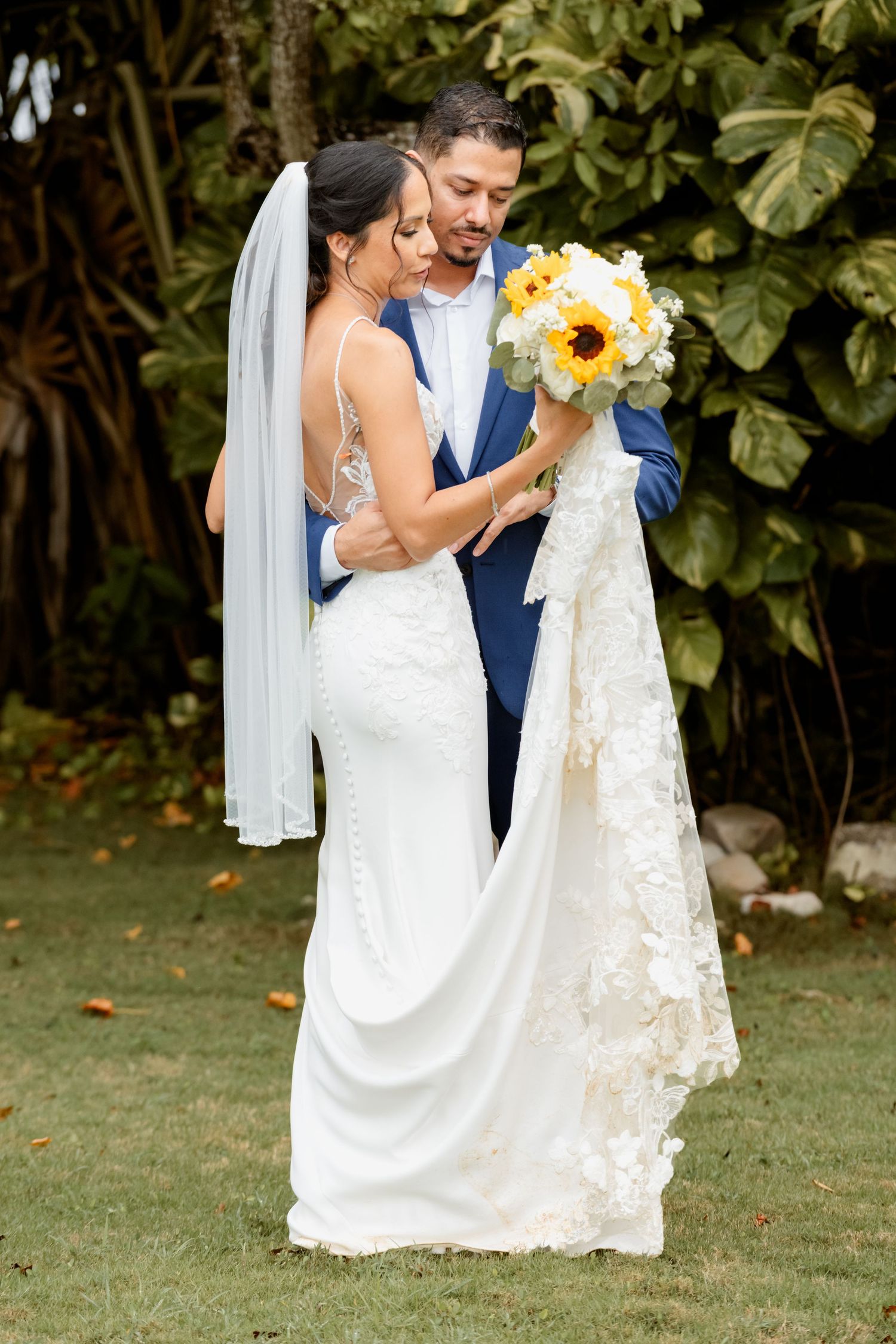 A bride in a white lace gown kneels outdoors with a young girl in a matching white dress holding sunflowers.
