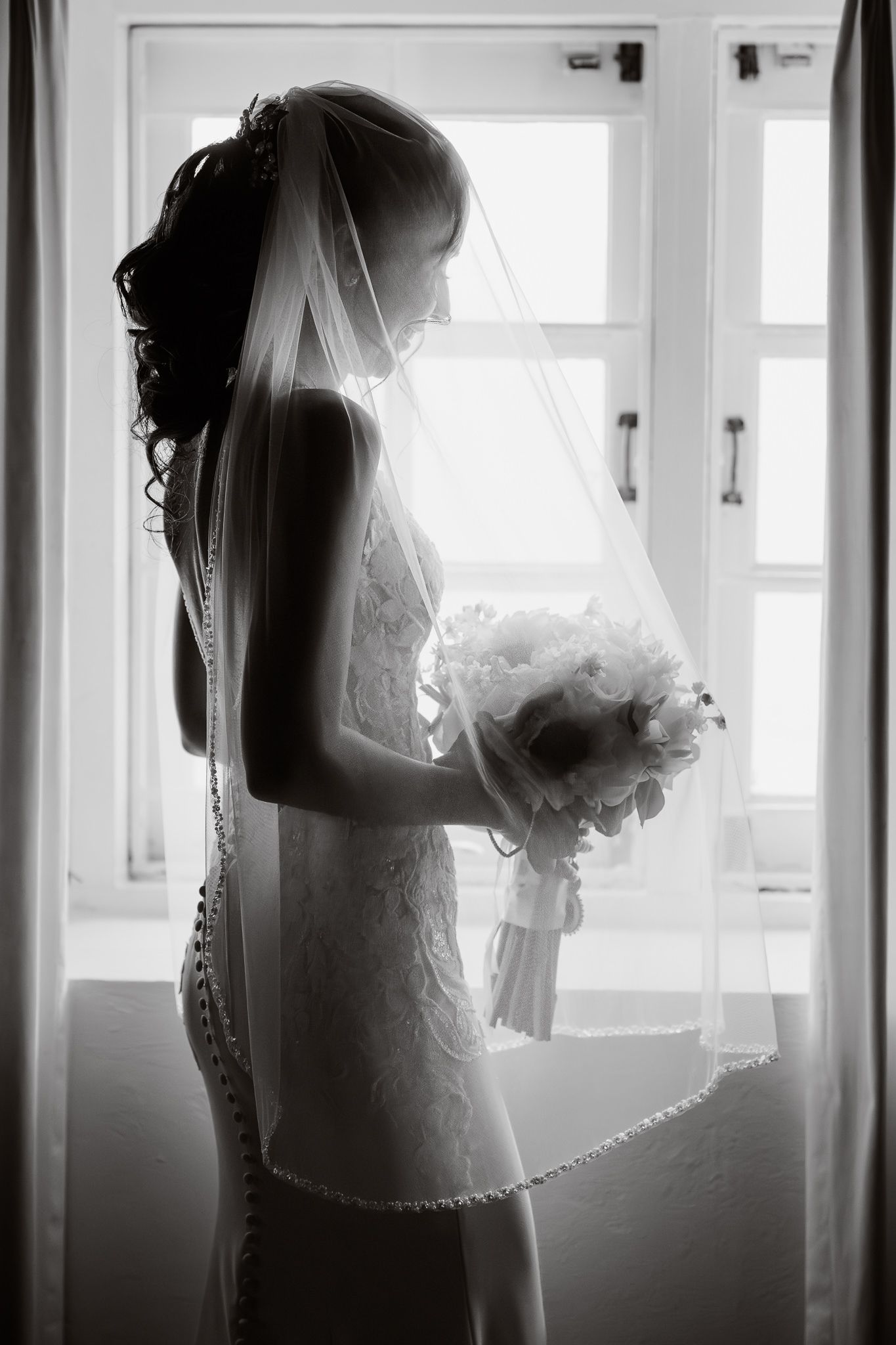 A silhouetted bride in a flowing white dress looks out a bright window in black and white photography.