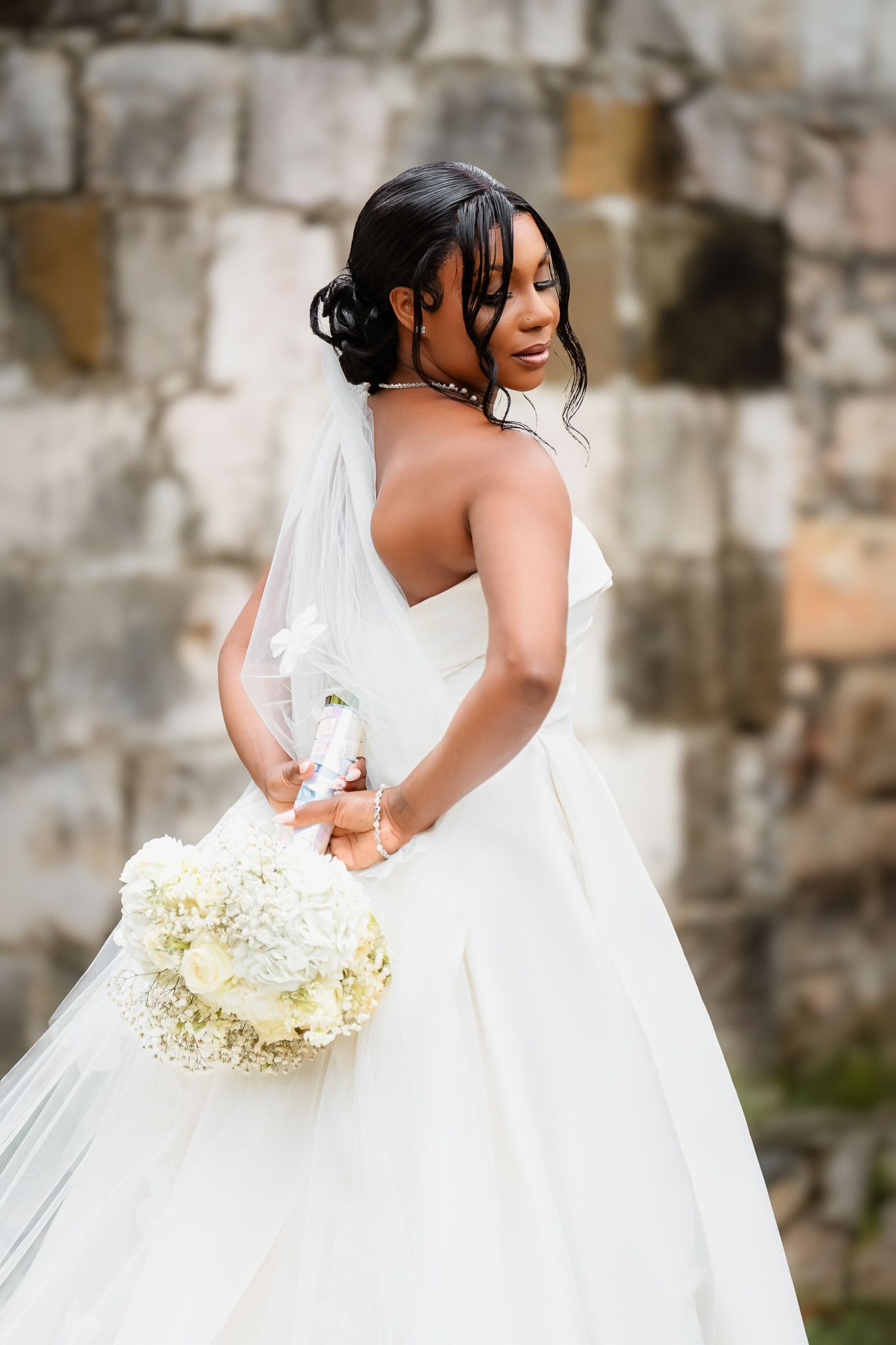 A Black bride in a strapless white ball gown holds a white floral bouquet, posing gracefully before a stone wall backdrop.