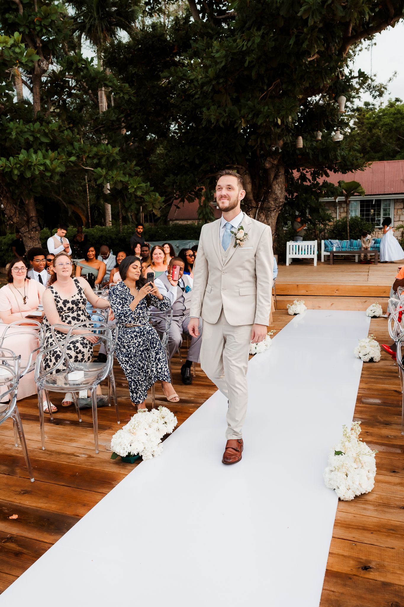 Groom in tan suit walks down white aisle runner at outdoor wedding ceremony with seated guests.