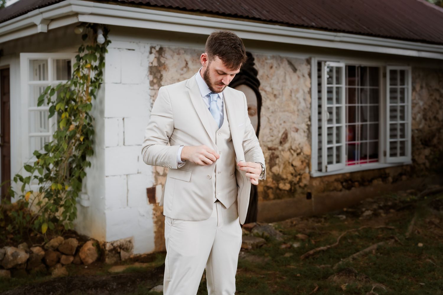 Groom in a beige suit adjusting his jacket outdoors in front of a rustic stone building on his wedding day.