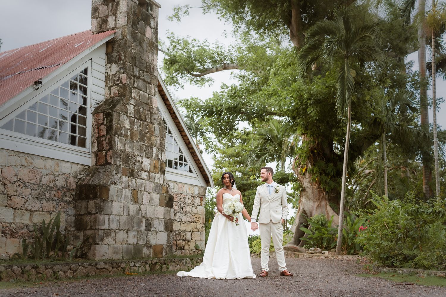 Bride and groom holding hands outside a rustic stone chapel surrounded by lush tropical trees on their wedding day.