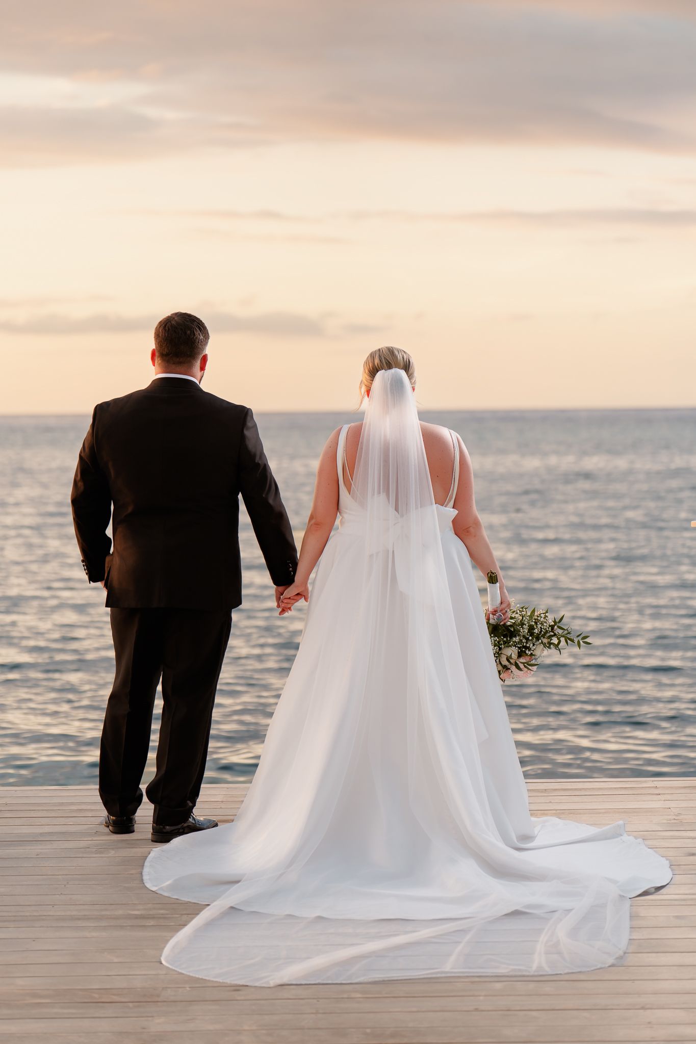 Bride and groom holding hands on a wooden dock overlooking a serene ocean at sunset, bride in white gown with veil.