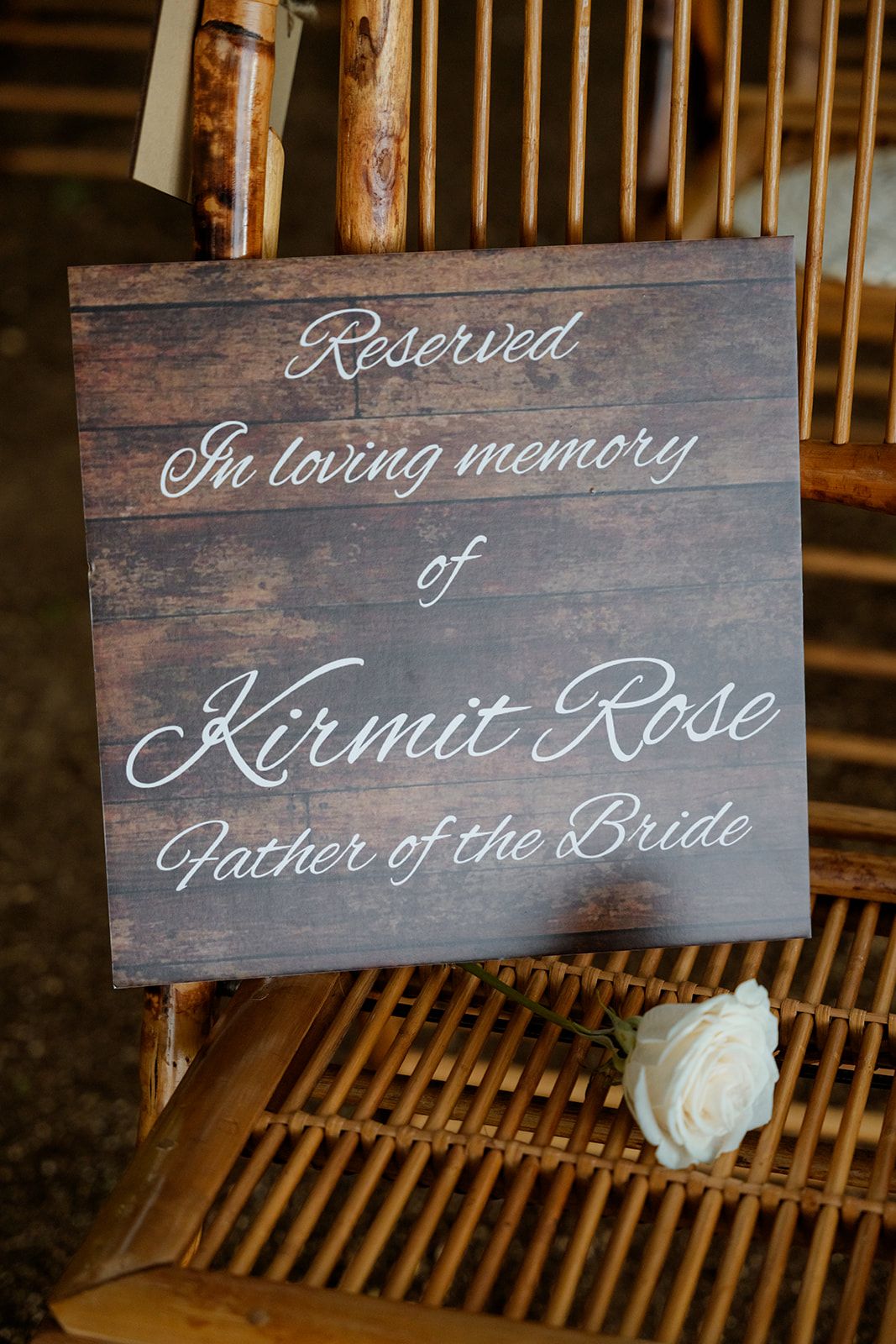 A wooden memorial sign reading 'Reserved in loving memory of Kermit Rose, Father of the Bride' with a white rose below.