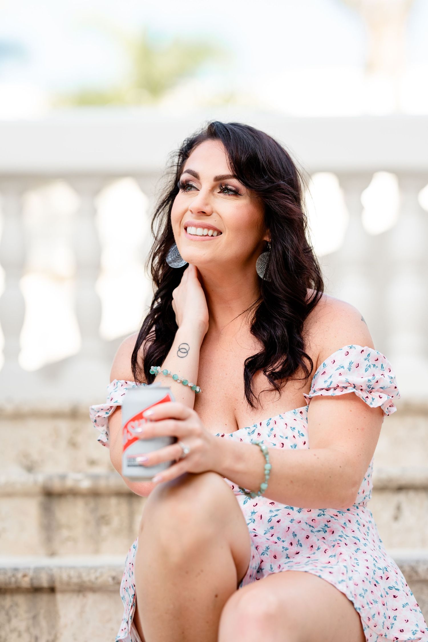 Woman in floral off-shoulder dress sits on stairs holding a cup while smiling outdoors on sunny day.