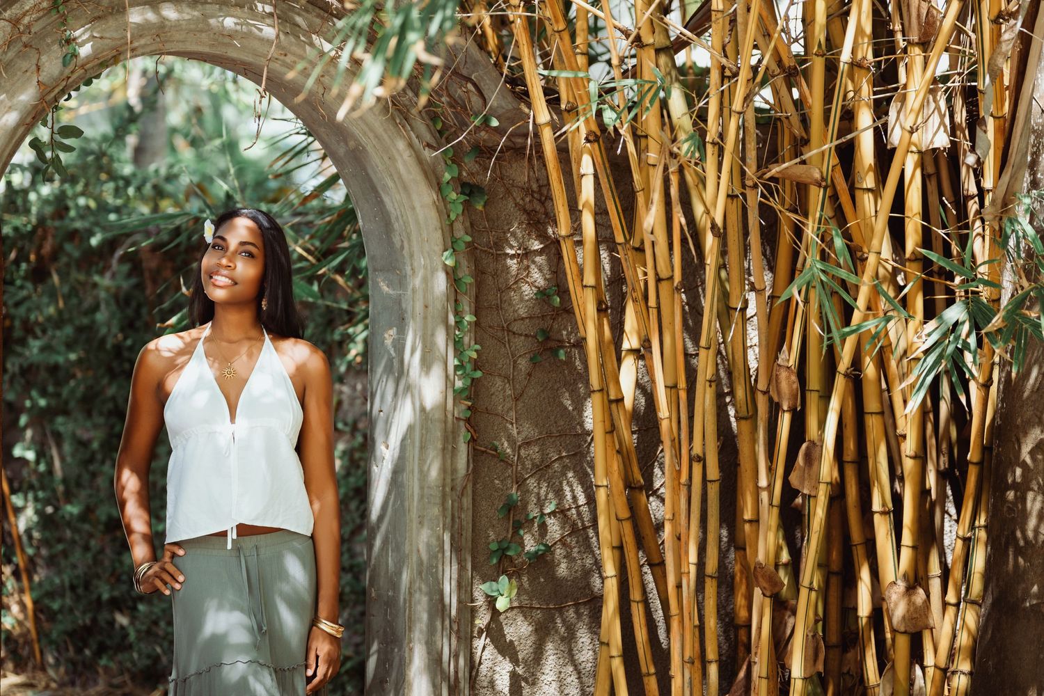 Woman in white dress poses elegantly among tall bamboo stalks in a lush garden setting.