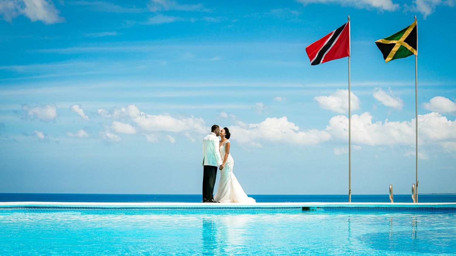 Couple kissing by infinity pool with Trinidad and Jamaica flags under blue sky at a destination wedding.
