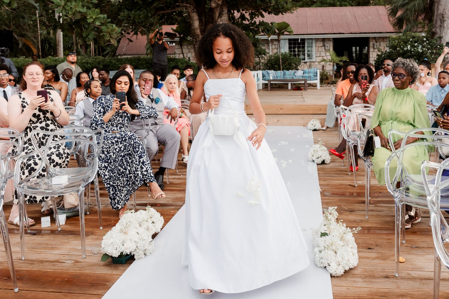 Young flower girl in white gown walks down outdoor wedding aisle with white floral arrangements and seated guests.