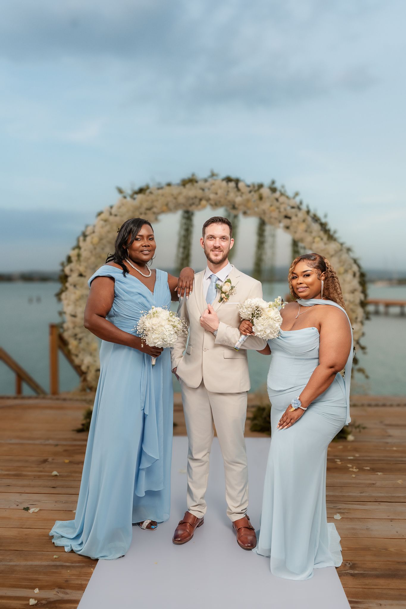 Groom in beige suit poses with two bridesmaids in blue dresses holding white bouquets at outdoor waterfront wedding.