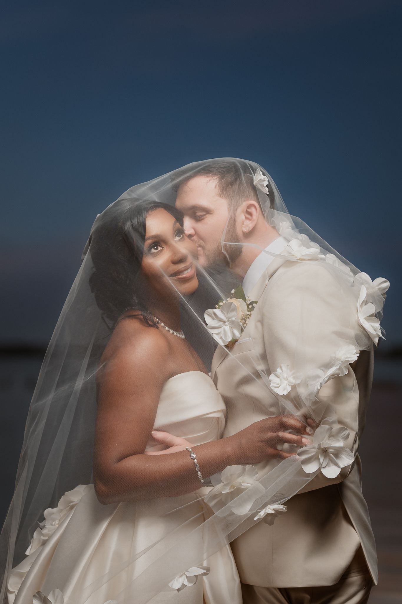 Interracial couple sharing a romantic moment under a floral veil at twilight, bride in strapless gown, groom in ivory suit.